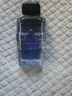 COPS 2.0 Eau de Toilette parfym - Unik parfymflaska formad som en polisfigur med blått glas och svart kork. COPS 2.0 Eau de Toilette har en cool och modern design som sticker ut i badrumshyllan. Perfekt för dig som gillar statement-flaskor och vill ha något annorlunda i din samling.