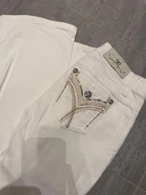 Vita bootcut low waist jeans från Miss Me - Snygga vita bootcut jeans från Miss Me med dekorativa detaljer och paljetter på bakfickan. Jeansen har låg midja, silverfärgade knappar och klassisk femficksdesign. Perfekta för dig som vill sticka ut med stilrena detaljer.