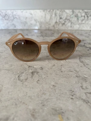 Beige runda Ray-Ban solglasögon - Snygga solglasögon från Ray-Ban med rundad form och beige bågar. Glasen är tonade i brunt och bågarna har en klassisk design med Ray-Ban-logga på sidan. Perfekta för att lyfta din stil under soliga dagar.