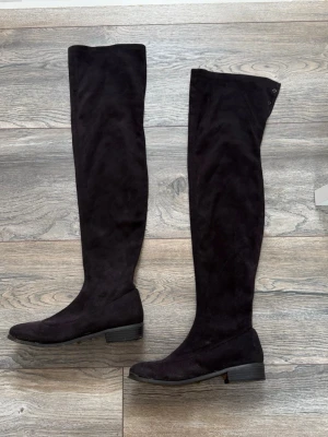 Svarta overknee stövlar i mocka - Säljer ett par svarta overknee stövlar i mjuk mocka-look med låg klack och rund tå. Stövlarna har en stilren design och små metallnitar upptill som detalj. Perfekta för att lyfta din outfit med en trendig touch. OBS att snöret inte finns med, utan du måste köpa nytt snöre för att kunna knyta skorna där uppe (se bild). OBS även att ena skon har en slitning på undersidan, se bild 