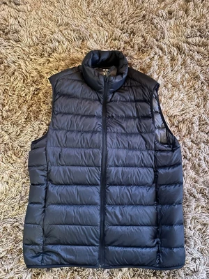 Svart dunväst från uniqlo - Snygg svart dunväst med quiltad design och hög krage. Västen har dragkedja framtill och är lätt vadderad för extra värme. Perfekt att slänga över en hoodie eller tröja för en chill look. Klassisk och enkel stil som funkar till det mesta.