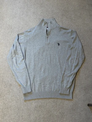 Halfzip U.S. Polo Assn. - Snygg ljusgrå tröja med halv dragkedja från U.S. Polo Assn. Tröjan har broderad logga på bröstet, ribbade muddar och hög krage. Perfekt till jeans eller chinos för en clean och stilren look.