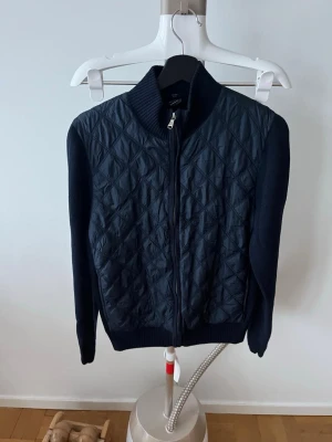 Boss cardigan - Snygg svart cardigan med quiltat mönster framtill och över axlarna. Dragkedja hela vägen, ribbad krage och muddar. Ärmar och rygg i mjukt stickat material, medan framsidan har en glansig quiltad yta. Perfekt för lager på lager och en clean look.