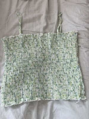 Blommig smockad linnetopp - Söt blommig linnetopp med smockad struktur och tunna axelband. Toppen har ett ljust grönt och vitt blommönster och är perfekt för varma dagar. Materialet känns lätt och luftigt, och modellen är croppad för en trendig look. Vet ej storleken