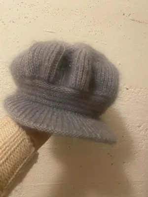 Grå stickad keps i mjukt garn - Säljer en grå stickad keps med mjuk och fluffig känsla. Kepsen har ribbstickad skärm och strukturstickat mönster upptill. Perfekt för dig som vill ha en unik och mysig accessoar till höst och vinter.