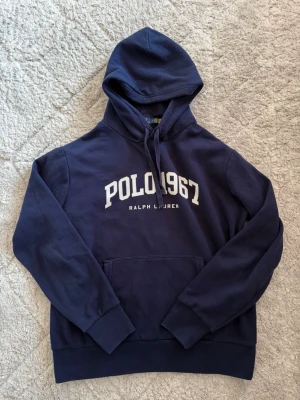 Marinblå hoodie Polo Ralph Lauren - Snygg marinblå hoodie från Polo Ralph Lauren med vit text 'POLO 1967' på bröstet. Klassisk känguruficka, huva med dragsko och ribbade muddar. Tillverkad i mjuk bomullsblandning som är perfekt för chill dagar. Priset är ej hugget i sten!