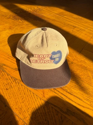Carhartt WIP beige och brun keps - Snygg keps från Carhartt WIP i beige med brun skärm. Framtill står det 'Heart of Detroit' i rött och vitt samt ett blått hjärta med Carhartt WIP-logga. Klassisk form med justerbar baksida och broderad ventilationshål. Perfekt för en avslappnad streetwear-look.