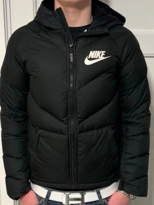 Svart dunjacka med huva från Nike - Snygg svart dunjacka från Nike med huva och dragkedja framtill. Jackan har två stora fickor och en vit Nike-logga på bröstet. Perfekt för kalla vinterdagar och ger en sportig look. Materialet är vadderat för extra värme.