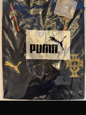 Svart Puma sporttröja Portugal - Helt ny! Snygg svart sporttröja från Puma med Portugals fotbollsförbunds logga i guld på bröstet. Tröjan har guldfärgad Puma-logga och detaljer i rött och grönt på axelbanden. Perfekt för fotbollsträning eller att bära till vardags.