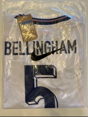 England fotbollströja 2026 - Helt ny! VM! Säljer en vit England fotbollströja från Nike med Bellingham och nummer 5 på ryggen. Tröjan har blå detaljer vid kragen och tryck i mörkblått. Perfekt för dig som vill stötta landslaget med stil.