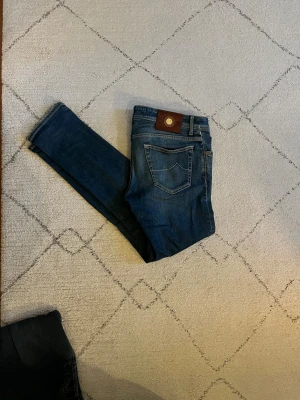 Jacob Cohen blå jeans style 622 - Snygga blå jeans från Jacob Cohen, modell style 622. Klassisk femficksdesign med knappgylf och detaljerad insida. Jeansen har en mörk tvätt och kontrastsömmar, samt ett brunt läderpatch bak i midjan. Perfekt för dig som gillar exklusiva jeans med unik känsla. Skick: 7-10 då jeansen är lagade vid midjan (bild 3) pris kan diskuteras vid snabb affär🤝