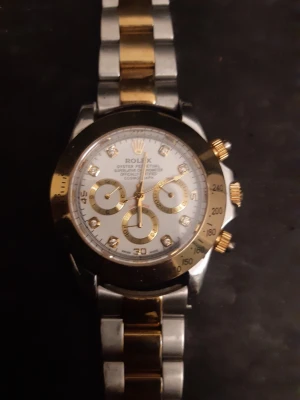 Rlx Daytona klocka stenar & guld detaljer  - Snygg Rolx klocka med tvåfärgad armlänk i guld och silver, lås saknas. Vit urtavla med gulddetaljer och stenar på index. Perfekt klocka för dig som gillar lyxig stil. Går som den ska men ny armlänk behövs. 