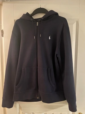 Mörkblå hoodie från Polo Ralph Lauren - Snygg mörkblå hoodie från Polo Ralph Lauren med dragkedja, två stora fickor framtill och klassisk huva med dragsko. Tröjan är sparsamt använd och har inga defekter och är köpt på Zalando!