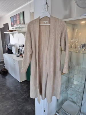 Beige lång kofta med knytskärp - Lång, ribbstickad beige kofta med öppen front och matchande knytskärp i midjan. Koftan har långa ärmar och en avslappnad passform. Materialet är mjukt och mysigt ut, vilket gör den superbekväm.