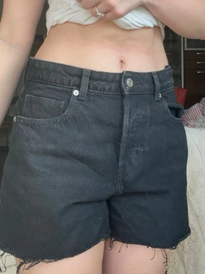 Svarta högmidjade jeansshorts - Svarta högmidjade jeansshorts med råa kanter och klassisk femficksdesign. Shortsen har bälteshällor och knappgylf. Perfekta för en avslappnad och trendig look.