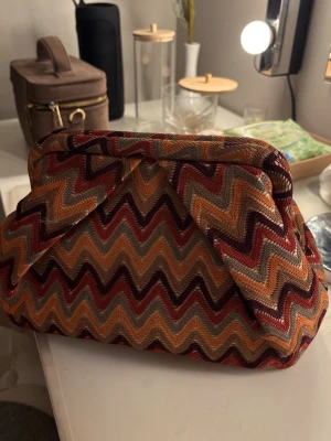 Mönstrad kuvertväska Beck Söndergaard - Snygg kuvertväska från Beck Söndergaard med färgglatt zickzackmönster i rött, orange, gult och beige. Väskan har en mjuk, vadderad form och är gjord i textilmaterial. Perfekt för att addera lite färg till din outfit.