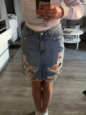 Blå jeanskjol med slitningar - Säljer en blå jeanskjol med råa, slitna detaljer och fransig nederkant. Kjolen har klassisk femficksdesign och knappgylf. Perfekt för dig som gillar en edgy och avslappnad stil.