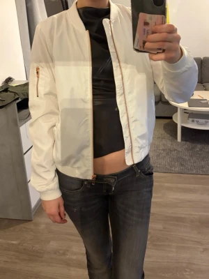 Vit bomberjacka från ONLY - Snygg vit bomberjacka från ONLY med klassisk passform och dragkedja i guld. Jackan har fickor på sidorna och en extra ficka med dragkedja på ärmen. Tillverkad i ett lätt och glansigt material som ger en fräsch look.