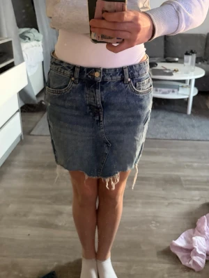 Blå jeanskjol med fransig kant - Säljer en klassisk blå jeanskjol med rå, fransig kant nertill och femficksmodell. Kjolen har normal passform och stängs med knapp och dragkedja framtill. Perfekt till en avslappnad look.