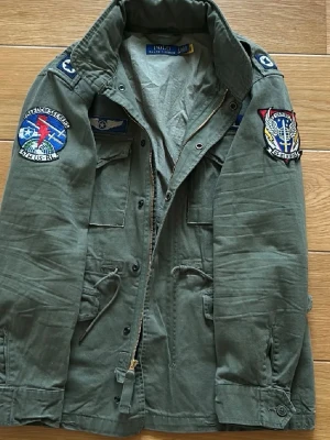 Grön vindjacka med patchar Polo Ralph Lauren - Snygg militärgrön vindjacka från Polo Ralph Lauren med flera coola patchar på ärmar och bröst. Jackan har dragkedja, knappar, fyra fickor framtill och justerbar midja med snörning. Perfekt för dig som gillar utility-stil och streetwear.