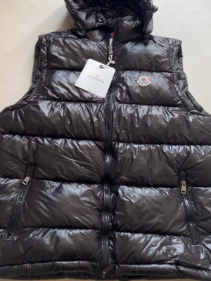Svart dunväst från Moncler med huva - Snygg svart dunväst från Moncler med glansig finish, huva och två dragkedjefickor framtill. Klassisk Moncler-logga på bröstet och dragkedja hela vägen upp. Perfekt för lager på lager och streetstyle.