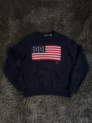 Mörkblå stickad tröja Polo Ralph Lauren - Mörkblå stickad tröja från Polo Ralph Lauren med amerikansk flagga och RL-broderi framtill. Tröjan har rund hals och ribbstickade muddar vid ärmslut och nederkant. Priset är absolut diskuterbart 