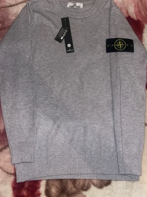 Grå stickad tröja från Stone Island - Snygg grå stickad tröja från Stone Island med klassisk logotyp-patch på vänster ärm. Tröjan har rund halsringning och ribbade muddar vid ärmslut och nederkant. Perfekt för dig som gillar stilrena och trendiga plagg. HAR INTE KVAR ETTUKETERNA
