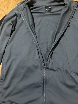 Svart träningsjacka från Under Armour - Säljer en svart träningsjacka från Under Armour med hel dragkedja och hög krage. Jackan är tillverkad i ett mjukt och stretchigt material som passar perfekt till träning eller sport. Diskret logga på bröstet och smal passform.