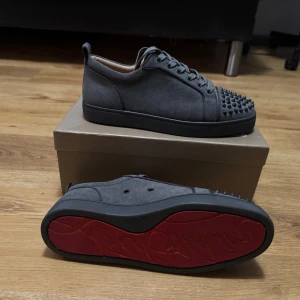 Christian Louboutin svarta sneakers med nitar - Säljer ett par svarta sneakers från Christian Louboutin med ikoniska röda sulor och coola svarta nitar på tån. Skorna är i mocka med snörning och har en stilren, rund tå. Perfekta för dig som vill sticka ut med en edgy vibe.