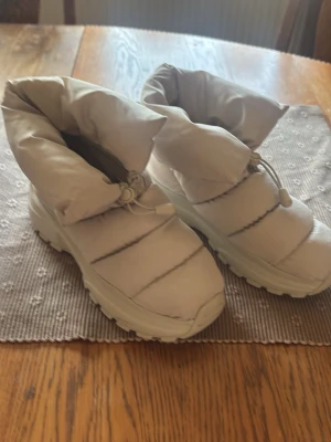 Beiga quiltade vinterkängor - Säljer ett par beiga, quiltade vinterkängor med tjock sula och dragsko upptill. Kängorna är vadderade och har en sportig, chunky design. Materialet är syntet och de är fodrade för extra värme under kalla dagar.