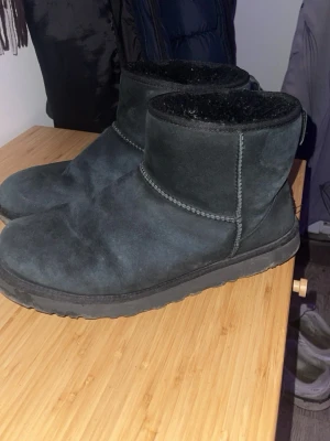 Svarta UGG boots i mocka - Svarta tofflor från UGG i mjuk mocka med varmt foder och låg skaft. Klassisk design med UGG-logga bak på hälen och robust sula. Perfekta för att hålla fötterna varma eller till chill-outfits.storlek 45 men passar lika bra från 43-45