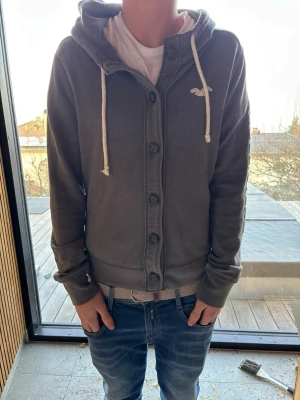Mörkgrå hoodie från Hollister - Mörkgrå hoodie från Hollister med knappar framtill, vit logga på bröstet och stora bokstäver 'HOLLISTER' längs ena ärmen. Utrustad med huva och vita dragsnören. Tillverkad i mjukt sweatshirtmaterial, perfekt för en avslappnad stil. Storlek m men har krympt till xs