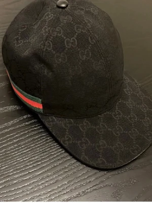 Svart keps från Gucci med GG-mönster - Säljer en svart keps från Gucci med det klassiska GG-monogrammet över hela kepsen. På sidan finns en grön och röd rand som ger en ikonisk touch. Kepsen är tillverkad i ett slitstarkt tyg och har en böjd skärm. Perfekt för dig som vill ha en stilren accessoar.