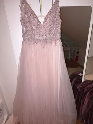 Ljusrosa balklänning  - Elegant ljusrosa långklänning med v-ringad spetsöverdel dekorerad med paljetter och pärlor. Kjolen är i flera lager tunn tyll som ger ett drömmigt fall. Perfekt för bal eller speciella tillfällen.
