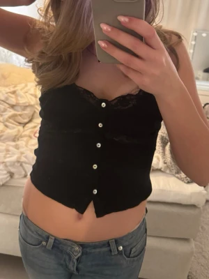 Svart ribbad linnetopp med spets - Svart croppad linnetopp från Zara med tunna axelband, spetsdetaljer vid bysten och knappar framtill. Toppen är ribbad och har en feminin v-ringning. Perfekt för en trendig och avslappnad stil. Säljer då den är för liten💓💓😙🥲