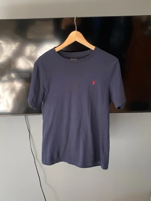 Mörkblå t-shirt från Polo Ralph Lauren - Mörkblå t-shirt från Polo Ralph Lauren med klassisk passform och korta ärmar. T-shirten har en liten röd broderad logga på bröstet och är tillverkad i mjuk bomull. Perfekt för en clean och stilren look. Storleken är 180 cm