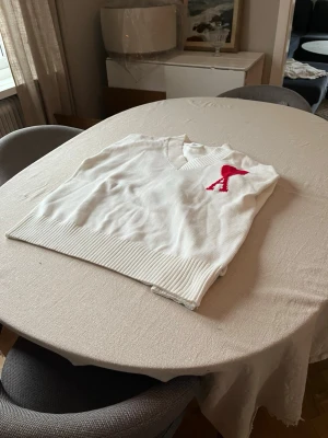 Vit stickad tröja från Ami Paris - Vit stickad tröja från Ami Paris med röd hjärtlogga och bokstaven A på bröstet. Tröjan har v-ringad hals, ribbade muddar och nederkant samt lång ärm. Perfekt för dig som gillar stilrena plagg med en twist.