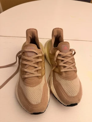 Beige Adidas Ultraboost sneakers - Snygga Adidas Ultraboost sneakers i beige och ljusbruna toner med rosa detaljer på tungan. Ovandelen är i ventilerande mesh och textil, med klassiska tre ränder i plast på sidorna och en tjock vit Boost-sula för extra komfort. Perfekta för en sportig och trendig look.