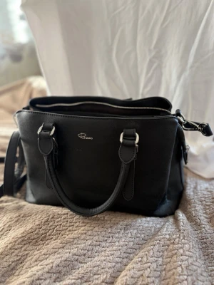 Svart handväska från Rizzo - Snygg svart handväska från Rizzo i stilren design. Väskan har två korta handtag, avtagbar axelrem och flera fack med dragkedja. Tillverkad i ett tåligt material med silverfärgade metalldetaljer. Perfekt för dig som vill ha ordning och stil. Helt ny endast stått i en hylla ca 3 månader sen jag fick den 