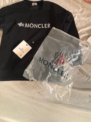 Svart sweatshirt från Moncler - Stilren svart sweatshirt från Moncler med stor vit logotyp broderad på bröstet. Tröjan har rund halsringning och ribbade muddar vid ärmslut och nederkant. Perfekt för dig som gillar exklusiva streetwear-plagg.