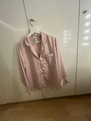 Ljusrosa pyjamaströja från H&M - Mysig ljusrosa pyjamaströja från H&M i mjukt, glansigt material. Klassisk skjortmodell med krage, knappstängning framtill och en bröstficka. Perfekt för chillkvällar hemma.