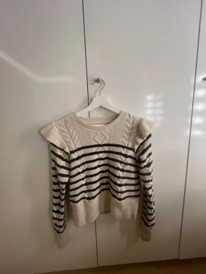 Randig stickad tröja med volang - Krämvit stickad tröja med svarta horisontella ränder och volangdetaljer vid axlarna. Tröjan har kabelstickat mönster framtill och baktill samt ribbade muddar vid ärmslut och nederkant. Rund halsringning och croppad passform.