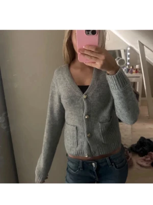 Grå kofta med knappar - Mysig grå kofta med v-ringning och guldiga knappar framtill. Koftan har två fickor och är stickad i ett mjukt material. Perfekt att slänga på sig över en topp för en chill och stilren look.