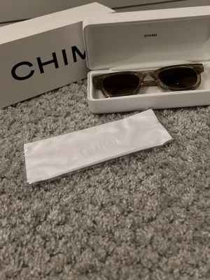 Ecru solglasögon från CHIMI - Snygga solglasögon från CHIMI som jag aldrig använt med transparent beige båge och mörka glas. Klassisk rektangulär form som ger en modern vibe. Kommer med originalfodral och putsduk. Perfekta för soliga dagar och en stilren look.