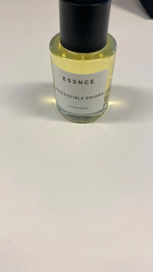Essnce Irresistible Enigma parfym - En stilren parfym från Essnce med namnet Irresistible Enigma. Flaskan är rund i genomskinligt glas med svart lock och innehåller en ljusgul vätska. Har använt den cirka två gånger 