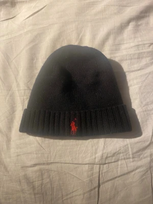 Svart mössa från Polo Ralph Lauren - Säljer en svart mössa från Polo Ralph Lauren i 100% merinoull. Klassisk ribbad kant och ikoniska röda loggan broderad framtill. Perfekt för dig som vill ha en stilren och varm accessoar till hösten och vintern.