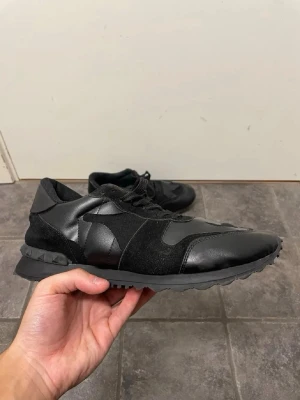 Valentino rock runners  - Snygga helsvarta rockrunners med både mocka och läderdetaljer. Skorna har en klassisk låg siluett, snörning och en robust yttersula. Perfekta för dig som gillar en stilren och clean look till vardagsoutfiten.