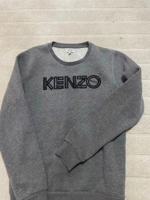 Grå sweatshirt från Kenzo - Säljer en stilren grå sweatshirt från Kenzo med svart broderad logga på bröstet. Tröjan har rund halsringning, långa ärmar och ribbade muddar vid ärmslut och nederkant. Perfekt för en avslappnad och trendig look.