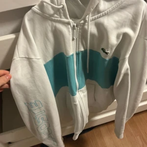 Vit hoodie med blå detalj från Lacoste - Säljer en vit zip-hoodie från Lacoste med blå vågig detalj över bröstet och stor Lacoste-logga i blått på ena ärmen. Klassisk krokodil-logga på bröstet och huva med dragsko. Perfekt för dig som gillar sportig och clean stil.