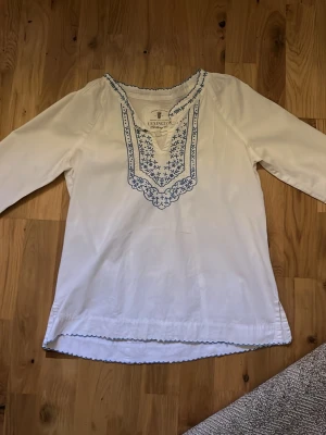 Vit blus med blå brodyr Lexington - Vit blus från Lexington med lång ärm och blå broderade detaljer runt halsringningen och nederkanten. Blusen har en avslappnad passform och är tillverkad i ett mjukt bomullstyg. Perfekt för en stilren och bohemisk look.